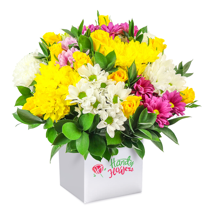 A densely packed mixed bouquet, shaped into a generous dome, fills a modern white square box that serves as both vase and gift container, all set against a pristine white background. The composition showcases a joyful blend of yellow, white and fuchsia blossoms, creating a vibrant focal point much like a burst of colour in a Stanmore garden at the height of summer. Large, full-headed yellow chrysanthemums form the visual anchor, their petals layered in countless soft curves that give the arrangement plush depth. Around them, clusters of yellow spray roses are placed at varying heights, some still tightly closed and others beginning to unfurl, contributing to the feeling of fresh, living growth. Balanced against these warm tones are crisp white flowers: small daisy-type chrysanthemums with bright green centres that bring an easy, casual charm, along with larger, fluffy white chrysanthemums that add volume and a cool contrast to the yellows. Striking fuchsia and magenta chrysanthemums are threaded throughout, some with lighter pink nuances, creating rich, jewel-like accents that stand out dramatically against both the whites and the yellows. Smaller pink and yellow rosebuds peek from between the larger blooms, intensifying the sense of abundance. Deep green, glossy foliage, likely a sturdy leaf such as ruscus, wraps the base and pokes between flowers, grounding the composition and enhancing the brightness of the petals. The white cube container has clean, simple lines, giving the piece a contemporary feel that would sit well in homes near Stanmore Golf Club or apartments around Canons Park. The bouquet appears freshly prepared and professionally arranged, ideal for local delivery as a birthday gift, a congratulations surprise, or a bright, uplifting arrangement for a dining table or office reception.