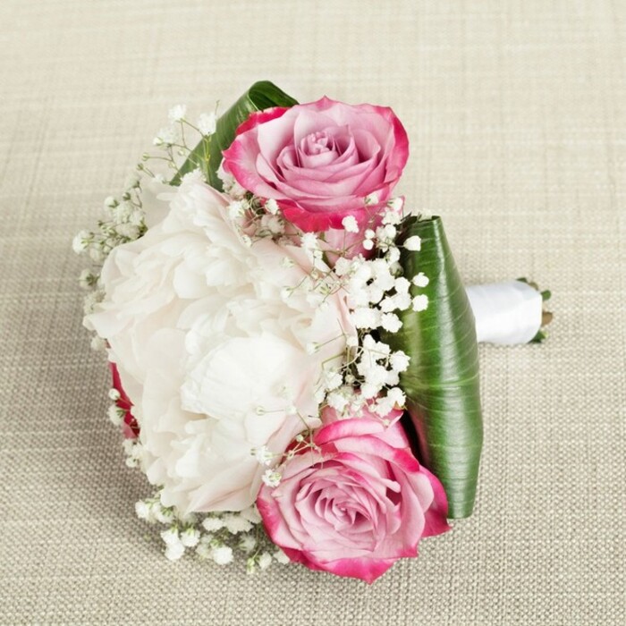Capturing the grace of a Stanmore celebration, this elegant bouquet is thoughtfully arranged on a natural linen background beneath soft, atmospheric lighting. Two pink roses, their petals painted in gradients from vivid magenta to tender blush and lavender, create focal points at the bouquet's corners. In between, a lush white peony unfurls, its ruffled blossoms full of delicate shadows and creamy freshness. Sprinkled through the arrangement are sprays of tiny baby's breath, introducing a cloud-like softness and ethereal movement. Encasing the blooms, broad, folded emerald leaves-perhaps aspidistra-form a bold, verdant frame, accentuating both structure and colour. The stems are neatly bound in a gleaming white satin ribbon, ready for someone's hand at a Stanmore wedding or a stroll through Bernays Gardens. Every petal and leaf catches the light, emphasizing craftsmanship, freshness, and subtle beauty. This bouquet from Flower Delivery Stanmore resonates with the tranquil spirit of the area, perfect for unforgettable occasions.