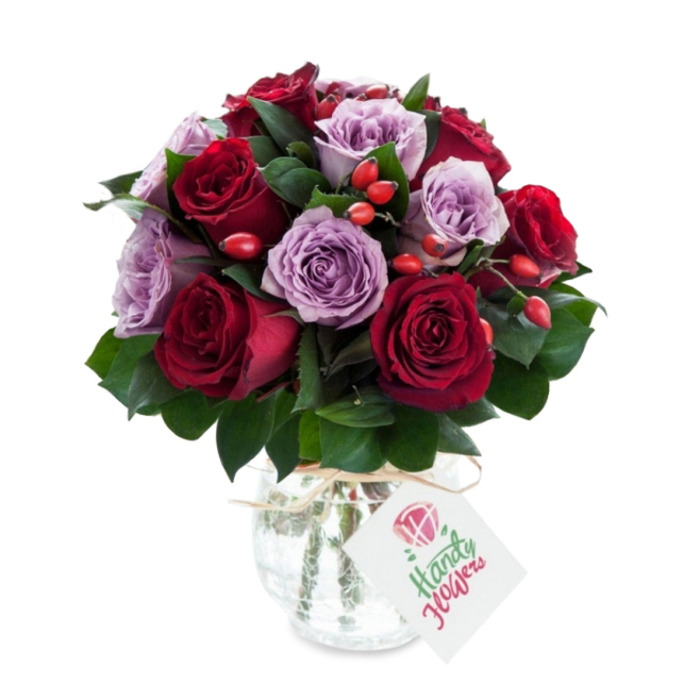 A richly hued bouquet that balances classic romance with a touch of rustic charm: densely arranged deep red and burgundy roses intermingled with soft lavender and lilac roses, all open and showing their layered centers. The composition is round and abundant, the darker red tones giving weight while the cooler mauves introduce a calming, sophisticated lift. Lush dark green foliage - likely Ruscus - sits beneath and between the blooms, its matt-to-gloss finish adding depth and shaping the silhouette. Sprinkled throughout are small clusters of red-orange rose hips that bring a seasonal, textural counterpoint; their glossy firmness and bright colour catch the eye amid the velvety petals. The bouquet rests in a rounded mercury-effect glass vase with a slightly tapered neck; the vase's silvered, crackled surface shimmers subtly, emphasising the blooms' colours, and a length of natural twine is tied at the throat to complete a handcrafted look. A simple white rectangular tag hangs from the twine, angled forward and showing a delicate botanical doodle rather than a logo, suggesting a personal note for the recipient. Photographed against pristine white, the arrangement reads as both luxurious and intimate. As a Stanmore florist I picture this delivered to a flat near Stanmore Station or brightening a table after a walk through Canons Park - Flower Delivery Stanmore can arrange local drop-offs that preserve the bouquet's freshness and presentation for anniversaries, birthdays, or quiet acts of kindness.