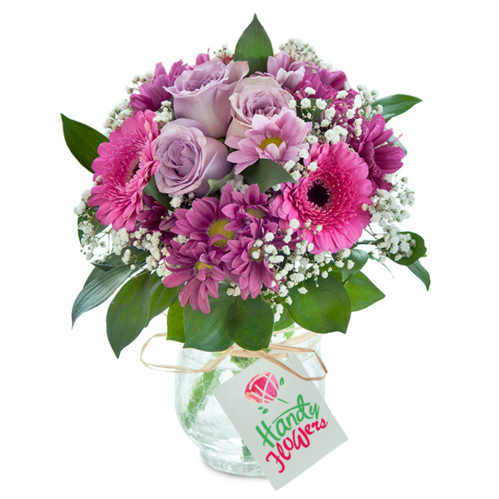 A magnificent, hand-assembled bouquet commands the spotlight against a pure white background, encapsulating the timeless elegance Stanmore residents cherish. At the heart, unfurling lavender roses impart a sense of serenity and gentle sophistication, their subtle fragrance reminiscent of tranquil afternoons by Stanmore's well-tended lawns. Surrounding them, radiant hot pink Gerbera daisies open boldly-each one's jet-dark center and vivid petals infusing the arrangement with cheerful vibrancy. Plush, deep magenta chrysanthemum-like blooms nestle between, creating a rich tapestry of shades and textures. Ethereal threads of white baby's breath are scattered throughout, brightening the bouquet and lending it an airy, cloud-strewn quality. Dark green foliage frames the composition, recalling the lush wooded edges of Stanmore Common. The whole bouquet is gathered in a sturdy, clear glass vase with a beautifully cracked texture, which refracts the light in subtle, sparkling patterns. A natural raffia bow encircles the neck, complete with a crisp white tag that awaits your message. Perfect for marking anniversaries, family celebrations, or heartfelt thank-yous, this arrangement brings a splash of artistry to homes and events across Stanmore, always guaranteeing freshness and exceptional quality.