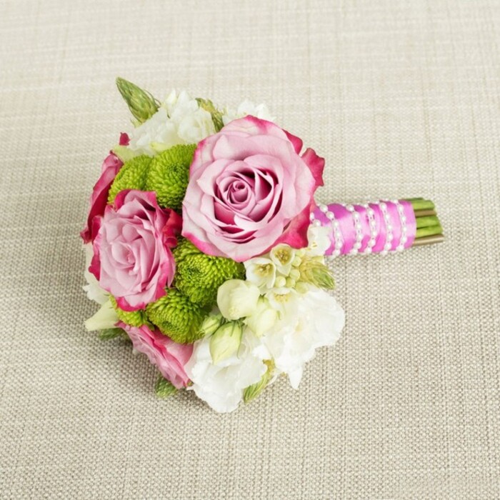 Displayed horizontally on a light beige woven fabric that could easily be a linen tablecloth laid out for a Stanmore afternoon tea, this small bouquet has the polished grace of a carefully considered wedding posy. At its centre, two prominent pink roses bloom fully, their petals forming tight yet generous spirals. The colour is soft at first glance-blush and shell pink-but deepens toward the outer edges into richer fuchsia and magenta tones, creating a natural gradient that adds depth and romance. A third rose nestles close by, slightly smaller but equally refined, reinforcing the sense of fullness without overwhelming the composition. Around the roses cluster vivid lime-green button chrysanthemums, each perfectly spherical head formed of tiny, densely packed florets that read as cheerful pom-poms. Their crisp green hue offers a refreshing contrast to the warm pinks, bringing the arrangement to life in a way that feels modern and playful. Cream and white filler flowers, reminiscent of petit lisianthus or freesia, are tucked between the bolder blooms, their slender, multi-layered petals giving a silky, almost whisper-light texture. Tiny star-like white blossoms, with delicate yellow centres, add detail that rewards a closer look, similar to spotting wildflowers along paths near Stanmore Country Park. The bouquet's hand-tied stems are neatly trimmed and wrapped in a rich fuchsia satin ribbon, whose smooth shine catches the soft, even light in the room. A slim strand of white faux pearls coils elegantly around the ribbon, lending an air of bridal sophistication. The neutral, textured background gently frames the flowers without distraction, allowing their colours and forms to stand out. This design would sit beautifully in the hands of a bridesmaid, serve as a petite bridal bouquet, or rest as a charming centrepiece at a local celebration in Stanmore, radiating freshness, intimacy, and carefully crafted style.