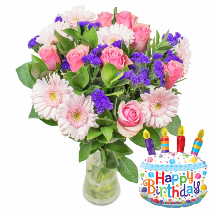 A vibrant, celebratory floral display set against a white backdrop, composed with thoughtful balance and hometown sensibility. At its heart are many soft pink roses, petals unfolding in layered spirals from pale blush to deeper pink centers, lending a classic, romantic core to the arrangement. Light pink Gerbera daisies sit among the roses, their broad, layered petals forming open disks with subtle yellow-green eyes that brighten the composition. Interspersed are abundant clusters of bright purple filler flowers, likely statice or limonium, whose small, papery blossoms create a textured contrast and enrich the colour palette. Delicate white sprays are woven through the arrangement to add points of brightness, while rich, glossy green foliage frames and supports the blooms, shaping the bouquet into a generous, natural silhouette. The flowers rest in a clear glass vase with a slightly wider base; the green stems and clear water are visible, underscoring the sense of freshness and honest craftsmanship. A metallic cake-shaped foil balloon sits to the right, decorated with white 'frosting', blue piped edging, multicolored sprinkles, and four striped candles in red, orange, blue and purple topped with tiny yellow flames. Bold, multicolored letters spell Happy Birthday across the front and a small KALEIDOSCOPE logo appears at the base of the balloon. This arrangement feels perfectly suited to a birthday table after a morning stroll through Canons Park or as a thoughtful surprise dropped off near Stanmore Broadway, reflecting a florist's attention to colour, scent, and conviviality in a familiar local setting.