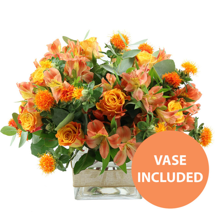 A richly coloured, seasonally inspired bouquet fills the frame, photographed against a bright white background so that each flower and leaf stands out in crisp detail. Dominating the arrangement are several beautifully formed orange roses, their petals unfurling in generous spirals that reveal subtle gradations of colour from deep tangerine at the outer edges to softer yellow tones towards the centre. Nestled beside them are rounded yellow garden roses, their tightly packed petals creating compact, rosette-like blooms that bring structure and a sense of classic elegance. Threaded through the arrangement are peach and light orange alstroemeria, or Peruvian lilies, with slim petals that open into star-like shapes; their inner surfaces are gently marked with fine streaks and dots, adding intricate visual interest. Punctuating the display are multiple small, spherical, spiky orange flower heads, similar to thistle or safflower, that introduce dynamic texture and a slightly rustic, meadow-like note. Encircling the flowers is a generous layer of foliage, including soft, silvery eucalyptus leaves and darker green fillers, which frame the bright blooms, provide contrast, and give the piece a full, abundant profile appropriate for a Stanmore family celebration. The stems are arranged neatly in a clear, square glass vase, the transparency revealing the straight green stems and clear water, suggesting recent, careful preparation by a local florist serving the Stanmore area around Canons Drive. Around the midsection of the vase, a band of natural beige burlap or jute is wrapped neatly, its fibrous, slightly rough texture bringing a warm, rustic charm that would suit a dining table in HA7 or a sideboard overlooking Stanmore Country Park. In the bottom right-hand corner of the picture, an eye-catching circular orange graphic contains the words VASE INCLUDED in bold white capital letters, clearly communicating that the container is provided. The bouquet conveys a mood of welcoming warmth and understated celebration, ideal for birthdays, get-well wishes, or simply brightening a quiet weekend at home.