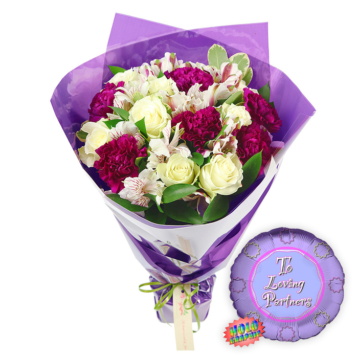 This image shows a romantic floral gift set laid out against a bright white background, as if ready for same-day delivery across Stanmore. The bouquet at the centre is lush and full, built around a cluster of creamy white roses, whose tightly furled petals form classic spirals that feel both timeless and refined. Nestled among them are numerous deep magenta to burgundy carnations, their ruffled petals layered thickly to create plush, cushiony blooms that introduce a sense of passion and richness. Woven throughout are stems of white alstroemeria, or Peruvian lilies, their elongated petals slightly flared and patterned with fine reddish-brown freckles and stripes on the inside, adding subtle detail that invites closer attention. Around this harmonious mix, fresh green foliage and hints of variegated leaves with light edges provide contrast, echoing the greenery of nearby Canons Park and softening the transition between flowers and wrapping.
The wrapping itself is meticulously done. Closest to the stems, a smooth sheet of white paper is folded diagonally, forming a clean, structured base that supports the flowers and accentuates their colours. Over this lies an outer layer of crinkled, translucent purple cellophane, which rises in loose folds and peaks around the bouquet, acting like a soft violet frame that diffuses the light and adds a gentle glow. At the point where the stems are gathered, a narrow bright green satin ribbon is tied into a tidy bow, its sheen catching the light; from it hangs a small rectangular light brown gift tag, suggestive of a personal note between partners. To the right of the bouquet, resting on the same white surface, is a round metallic purple foil balloon with a scalloped, rosette-style edge. The centre of the balloon is a paler lavender circle, printed with the words To Loving Partners in graceful pink script, while a tiny rainbow-coloured holographic sticker with jagged edges is fixed near the lower left, hinting at a special reflective finish. Together, this carefully coordinated bouquet and balloon create a heartfelt romantic gift, ideal for anniversaries, Valentine's surprises, or celebrating relationship milestones in Stanmore and surrounding areas.
