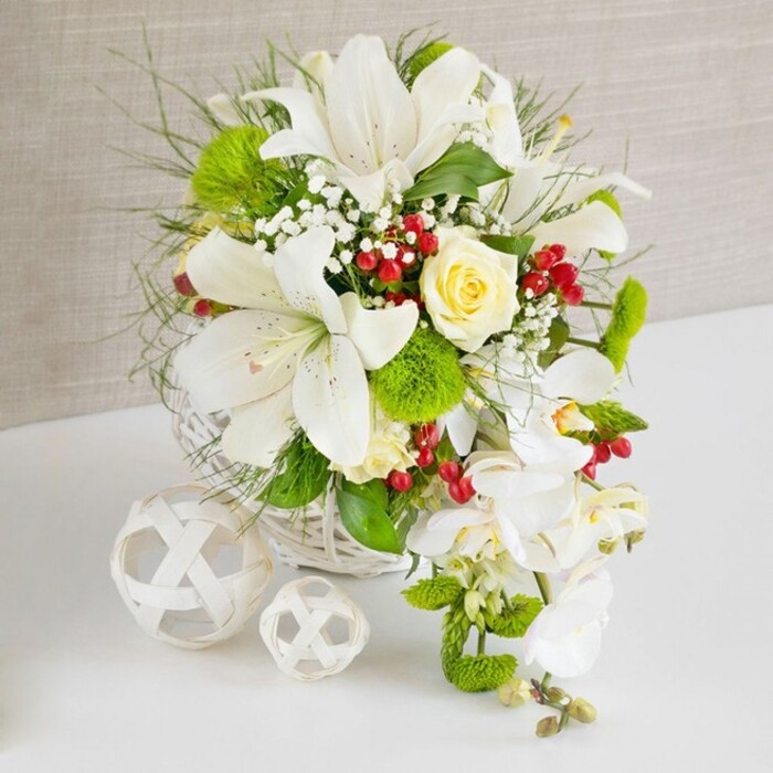 A meticulously arranged white and green centerpiece rests on a spotless white tabletop, its refined beauty perfectly suited to the tranquil, residential feel of Stanmore. The flowers are gathered into a low, round white basket crafted from woven wicker, the pale strands forming a soft circular frame. Rising from this base, several large white lilies open fully, their trumpet-shaped petals sweeping outward to reveal speckled throats and golden stamens, suggesting a gentle fragrance that would suit an intimate dining room overlooking Stanmore's tree-lined streets. Nestled close to the top centre, a single creamy ivory rose becomes the heart of the design, its tightly layered petals creating a sense of focus and romance. From the right-hand edge, a cascade of white phalaenopsis orchids flows downwards in a graceful curve, each orchid flower broad and smooth, creating a waterfall effect that brings movement and drama, much like a bridal bouquet carried through nearby Stanmore Country Park. Interwoven among these main flowers, bright green pom-pom dianthus add a cushiony, moss-like texture, while sprays of feathery greenery and broader leaves provide volume and shape. Tiny clouds of white gypsophila hover delicately between the stems, softening the transitions and enhancing the arrangement's airiness. Small clusters of ruby-red berries, glossy and round, appear at intervals, giving sparks of rich colour that feel festive yet controlled. To the left of the basket, two decorative white wicker balls, one larger and one smaller, rest on the table, their intricate latticework echoing the basket and lending a contemporary, styled note. The background is a soft, beige fabric with a subtle weave, allowing the crisp whites and vibrant greens to stand out in high contrast. The composition radiates purity, sophistication, and quiet joy, an ideal centerpiece for weddings, anniversaries, and special celebrations throughout Stanmore.