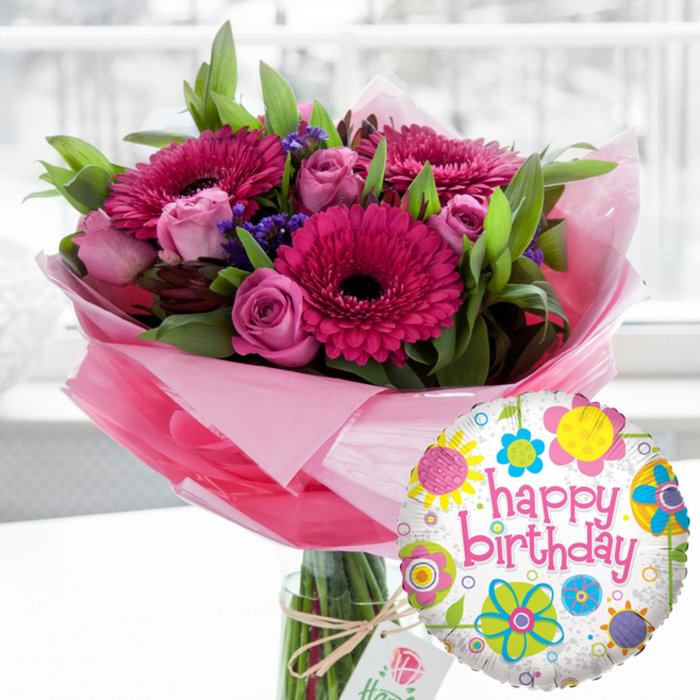 An elegant birthday scene unfolds around a luxurious pink bouquet and coordinating foil balloon, the kind of gift you might send from Flower Delivery Stanmore to brighten a loved one's home near Canons Drive. The flowers are arranged in a clear glass cylindrical vase, the stems neatly trimmed and immersed in water that glints in the soft indoor light. Dominating the display are several large gerbera daisies in saturated tones of fuchsia and magenta, their petals radiating in perfect concentric layers around dark, almost black centers that give the bouquet a confident, celebratory heart. Nestled among them are numerous blush and pastel pink roses, some still tightly furled and others just beginning to unfurl, revealing their intricate spiral of petals; they lend a sense of romance and refinement to the arrangement. Threaded throughout are clusters of tiny deep purple blooms, likely statice, providing a feathery texture and a cool contrast to the warmer pink hues. Lush green foliage, with smooth, slightly glossy leaves, frames the flowers and adds volume, suggesting a freshly gathered garden feel. Around the base of the flowers, soft, translucent pink cellophane is wrapped and lightly crinkled, creating a delicate, shimmering ruffle that softens the transition into the vase. Tied around the neck with rustic twine hangs an off-white rectangular gift tag adorned with a subtle rose emblem, offering a quiet nod to quality and attention to detail. In the foreground to the right, a round Mylar balloon gleams, its white surface dotted with grey speckles and ringed with playful floral motifs in bright yellow, sky blue, lime green, and vivid pink. Bold pink script spells out happy birthday, making the purpose of the gift unmistakable. Behind it all, the background melts into a gentle blur suggestive of a sunlit Stanmore living room, allowing the flowers and balloon to command all attention and conveying the joy, affection, and festivity of a milestone birthday.