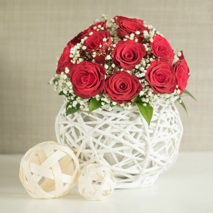 The image reveals a beautifully balanced floral centrepiece, perfectly suited to a wedding table in Stanmore, perhaps set near Belmont Circle or along Marsh Lane. At the heart of the composition, a generous cluster of deep red roses forms a rounded, almost spherical bouquet. Each rose is at its best, fully opened yet still structured, with velvety petals spiralling tightly at the centre and gradually loosening towards the edges. The roses are grouped closely, creating a rich, opulent texture that draws the eye immediately. Interlaced among the blooms are sprays of delicate white baby's breath, each tiny blossom adding softness and a subtle, cloud-like effect, lightening the mood of the arrangement. Small, tasteful hints of green foliage are visible where the stems gather, introducing a gentle touch of nature's freshness without overwhelming the roses. The bouquet is nestled into a distinctive white vase with a bulbous shape and an airy, open-weave design, as if slender white twigs or rattan strands have been woven together into a hollow globe. This intricate lattice casts fine shadows on the smooth, pale surface beneath, lending depth and texture to the scene. To the left side of the vase, two decorative orbs made of light, woven material in soft cream or beige tones rest on the table. The larger ball sits in front, the smaller one just behind, their open lattice designs echoing the pattern of the vase and adding visual balance. The background is a softly textured, neutral wall, slightly blurred, which allows the bold reds and crisp whites to stand out clearly. Overall, the scene conveys romance, elegance, and careful design, ideal for marking an anniversary, engagement, or intimate wedding celebration in Stanmore.