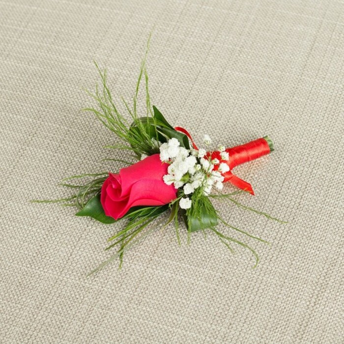 A finely detailed boutonnière featuring a single, tightly furled fuchsia rosebud is displayed at a gentle diagonal across a pale beige fabric that looks like textured linen, giving the scene a soft, natural backdrop. The rose, with its compact spiral of velvety petals, glows in shades of deep pink and magenta, the outer petals slightly lighter and the inner ones rich and saturated, drawing the eye instantly to its heart. Nestled closely around the rose are airy clusters of white baby's breath, each cluster made up of many tiny, star-shaped blossoms that lend a cloud-like delicacy and soften the intensity of the central bloom. Emerging behind and around the flowers is a mix of vibrant green foliage: some fronds are fine and feather-like, splaying outward in graceful arcs, while a few broader, darker leaves sit nearer the base, anchoring the design and giving it structure. All the stems are carefully gathered and wrapped with a vivid red satin ribbon, the ribbon wound tightly and evenly to create a sleek handle, ending with a small decorative loop that hints at the florist's precise craftsmanship. The light in the image is bright yet gentle, with no harsh shadows, allowing the colours and textures to be clearly seen-the smooth rose petals, the slightly matte baby's breath, the glossy ribbon. The overall impression is of a freshly made boutonnière from a trusted Stanmore florist, perhaps prepared at Flower Delivery Stanmore for a groom heading to a ceremony near Stanmore Hill or a formal event by Bentley Priory. It feels timeless and romantic, a classic lapel flower that combines traditional elegance with a vibrant, modern twist on pink and red tones.