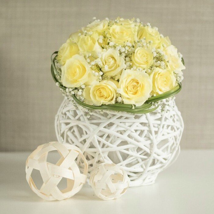 Captured in soft, diffused light, this floral centerpiece feels like it belongs at the centre of a beautifully dressed table in a Stanmore hotel or private home. The arrangement features a rounded dome of soft yellow roses, closely packed so that their velvety petals form a continuous blanket of colour, with each bloom unfurling in concentric whorls. The shade of yellow is gentle and creamy, more like melted butter than bright sunshine, lending the bouquet a calm, sophisticated character. Nestled between the roses are clusters of white baby's breath, their tiny blossoms scattered along fine, branching stems, adding lightness and texture, and making the arrangement seem almost cloud-like from a distance. A narrow band of smooth, dark green foliage encircles the base of the blooms, providing a clear line before the eye meets the vase and enhancing the sense of craftsmanship. The vase itself is a striking decorative piece: white and rounded, formed from slender, interwoven strands that create an open, basket-like lattice. This airy construction casts gentle shadows onto the glossy white tabletop, echoing the woven patterns of natural rattan. Just in front of the vase lie two decorative spheres crafted from pale, natural material, also open and woven, their sizes slightly different for visual interest. The background is a soft, neutral beige with a hint of fabric-like texture, subtly blurred, as if the scene were set in a quiet dining room or reception space off Marsh Lane or near Stanmore Country Park. The image conveys a sense of poised celebration and thoughtful design, ideal for wedding table arrangements, engagement dinners, or refined floral delivery across Stanmore.