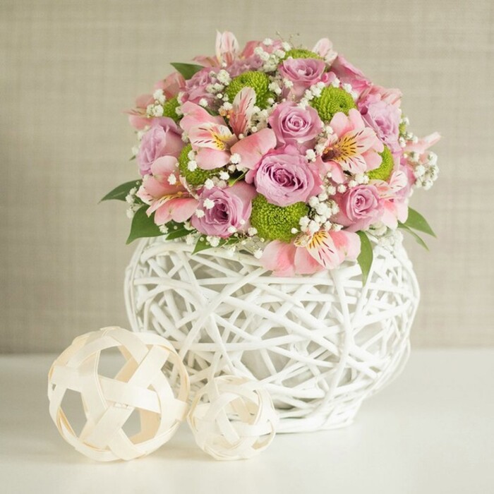Taking center stage in a tranquil Stanmore interior, this exuberant floral arrangement combines tradition and contemporary flair. Nestled within an intricate white vase-openly woven from slender bands to create a sturdy, rounded shape-lush blooms rise in layered profusion. The bouquet is alive with pastel pink roses and lavender blossom, petals velvety and open, accented by airy drifts of baby's breath, their tiny white flowers providing a soft haze between larger blooms. Elegant pink alstroemeria, their slender petals etched with fine stripes and dappled with yellow or deeper pink, weave through the composition alongside playful, spherical lime-green button chrysanthemums for a modern accent. Rich, dark green leaves peek from below, grounding the pastel colors and bringing echoes of the nearby Bentley Priory nature reserve. Two hand-crafted decorative spheres-fashioned from pale woven strips in natural tones-are artfully set to the left of the vase on a gleaming white surface, their openwork mirroring the artistry of Stanmore's creative community. The subtle, textured beige background allows the softness of each bloom and the crisp contemporary lines to shine. Whether destined for a wedding table, elegant home setting, or delivered to a loved one in Stanmore, the arrangement embodies freshness, celebration, and the unique character of this northwest London enclave.