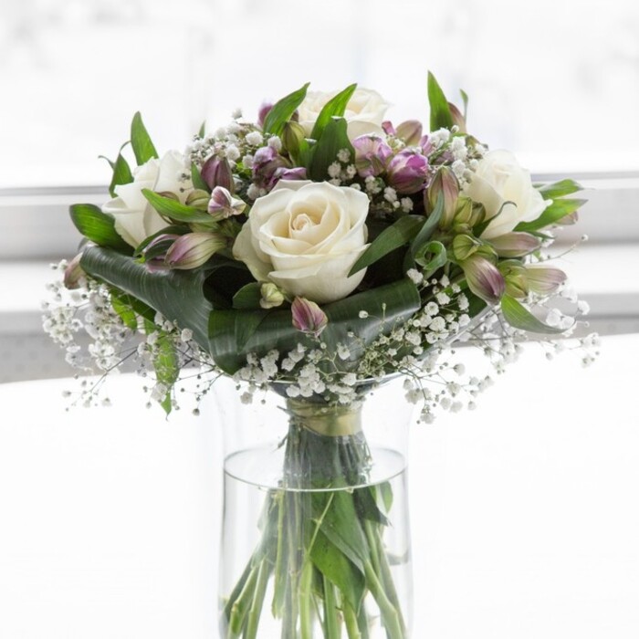 In this graceful bouquet, creamy white roses and soft lavender alstroemeria are gathered in a clear cylindrical glass vase, suffused with gentle daylight that feels like it is filtering through a clean window in a Stanmore townhouse. The white roses are fully open yet still crisp, their layered petals unfolding like silk ribbons into rounded, romantic blooms that instantly draw the eye. Interwoven with them are alstroemeria blossoms in nuanced tones of lilac and muted purple, some flowers fully revealed with delicate darker streaks along their petals, others still held in slender buds wrapped in fresh green sepals. Between these focal flowers, clusters of tiny white baby's breath float like miniature clouds, filling any gaps and lending the whole design a delicate, misty softness. Around the edge and peeking through the centre, lush foliage in deep and mid-green shades provides contrast and structure, with glossy leaves that catch the light and thinner stems that add a sense of natural movement. The stems of all the flowers are clearly visible through the transparent vase, trimmed neatly, standing in clear water that reflects a commitment to freshness and care, akin to something handpicked from gardens near Stanmore Country Park. The arrangement is thoughtfully rounded and full, with no harsh lines, creating a calm, balanced silhouette that feels both classic and contemporary. The softly diffused background keeps the focus on the flowers, while suggesting a peaceful interior setting-perhaps a dining table in a Stanmore home ready for a family gathering, a reception desk near Canons Park station, or a bedside in need of comfort. The bouquet's gentle white and lavender palette conveys purity, remembrance, and quiet joy, making it well-suited for heartfelt sympathy, milestone celebrations, or simple moments of everyday beauty across the local HA7 community.