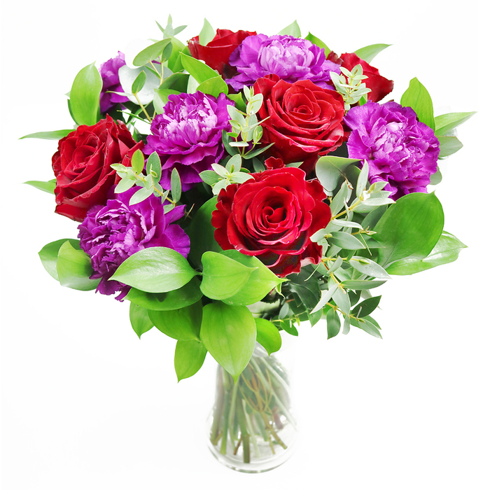 This image presents a lush and polished bouquet of red roses and purple peonies arranged in a clear glass vase, exuding both modern elegance and timeless romance. The red roses dominate the upper part of the arrangement, their petals thick and velvety, curling back in generous spirals that reveal a deep, saturated shade of crimson. Intermingled with them are large, plush purple peonies whose many layered petals form rounded, cloud-like blooms; the ruffles catch the light in subtle highlights, giving a sense of depth and movement. Around the main flowers, a ring of greenery fans out: smooth, glossy leaves create a rich, green frame, while slender stems of eucalyptus weave through the composition. The eucalyptus leaves, with their soft silver-green colour and gently curved shapes, contribute a relaxed, contemporary feel and suggest a cool, slightly herbal scent. The glass vase is simple and cylindrical, allowing the stems to be seen clearly-trimmed neatly and criss-crossing slightly, they give the design a grounded, intentional structure. Soft daylight appears to wash over the bouquet, much like the light you might find in a Stanmore living room with views towards the open spaces of Stanmore Country Park, intensifying the deep reds and bringing a regal richness to the purples. One can almost imagine the bouquet placed at the centre of a celebratory table, its gentle fragrance mingling with the atmosphere of a family gathering or romantic dinner. Crafted with the precision of a local florist, this arrangement feels ideal for marking anniversaries, engagements, or heartfelt congratulations delivered in and around Stanmore, from quiet residential avenues off Marsh Lane to elegant homes near Stanmore Golf Club.
