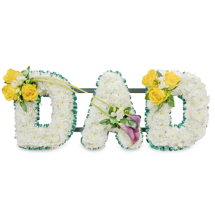 A carefully crafted memorial display spelling the word DAD in three large, freestanding capital letters, each letter entirely composed of compact clusters of pristine white chrysanthemums that form a soft, pillowy surface. The letters are edged with a ruffled, deep-green satin ribbon that provides a clean, contrasting outline against the white floral mass, and thin dark-green metal support bars run horizontally behind the letters to bind them into a single, cohesive tribute. The first letter, the left D, is embellished on its upper left curve with a bright, sunlit cluster of approximately three to four yellow roses, accompanied by delicate white filler flowers-freesias or gypsophila-and fresh green foliage; two long, bare green stems sweep diagonally from this cluster toward the centre, creating a gentle gesture. The centre A carries a distinctive, refined accent low on its left leg: a pair of striking deep purple or burgundy calla lilies whose trumpet shapes offer an elegant counterpoint to the rounded chrysanthemums, neatly surrounded by tiny white accents and glossy greens. The right D mirrors the left with a matching yellow-rose cluster positioned on its upper right curve, balancing the composition. Set against a plain white backdrop in soft, diffuse light, the arrangement reads as both stately and tender-white symbolising peace, yellow evoking warmth and remembrance, and purple conveying reverence. This bespoke piece is appropriate for funerals and memorials throughout Stanmore and nearby neighbourhoods such as Canons Park and Harrow, and it reflects the quiet care of a local florist arranging flowers with attention to texture, scent, and dignified presentation.