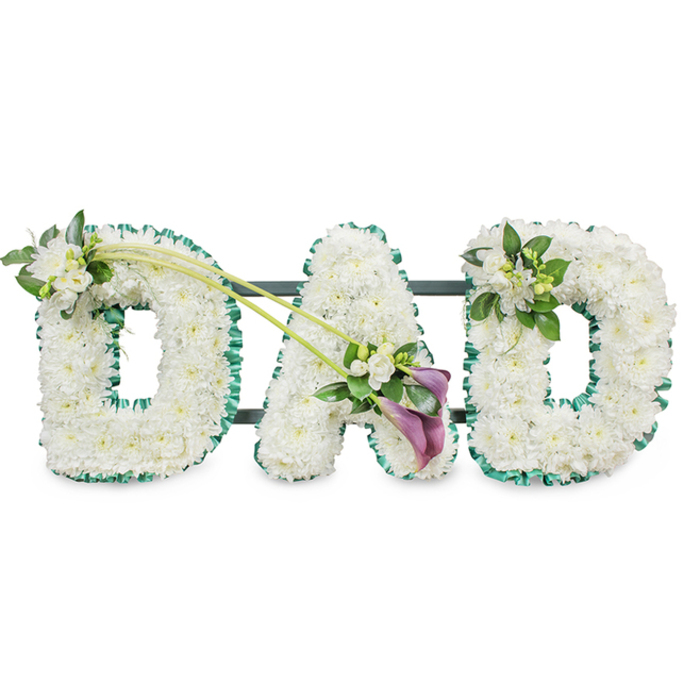 This photograph shows an elegant DAD funeral flower tribute created with the kind of meticulous care you might expect from experienced florists serving Stanmore and the nearby residential streets off Marsh Lane and Uxbridge Road. The three letters, D, A, and D, stand upright in a neat row against a pure white background, making every detail of the design easy to see. Each letter is crafted as a three-dimensional block and entirely covered with masses of pristine white chrysanthemums or similar small, rounded blooms. The petals overlap to form a dense, cushion-like surface, giving a sense of softness and serenity that suits a final farewell to a father. A rich emerald green satin ribbon runs around the entire perimeter of every letter, ruffled into gentle waves. This ribbon adds definition and frames the white flowers, its fresh green tone evoking the parkland around Bentley Priory and the tree-lined streets of Stanmore. Behind the blooms, slender dark bars hint at the supporting frame that keeps the tribute standing during the service.
Extra floral details lend character and emotional nuance. On the top left curve of the first D, a delicate spray of white freesia and lush green foliage is nestled into the chrysanthemum base, the freesias rising above with their elegant buds and open flowers. From this point, two long, graceful green stems arch diagonally across toward the central A, creating a gentle line of movement across the tribute. At the meeting point on the A, an eye-catching spray features two deep purple calla lilies, their velvety trumpet shapes curling inward, surrounded by more white freesia blossoms and glossy leaves. This deeper shade of purple adds depth and solemn beauty to the composition. The final D is decorated at its upper right side with a smaller matching cluster of white flowers and greenery, tying the design together. The overall effect is one of calm dignity and heartfelt remembrance, a traditional yet deeply personal DAD tribute that families in Stanmore might choose to express love, gratitude, and lasting memories.