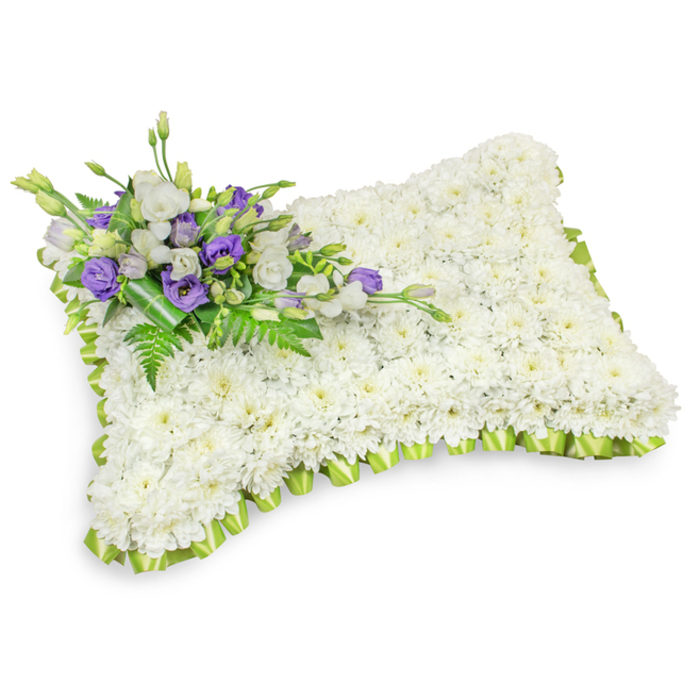 An exquisitely made sympathy pillow, its surface completely covered with a dense field of small white chrysanthemum blooms that read as a smooth, serene plane. The piece is shaped as a rectangular cushion with softly rounded corners and a slightly domed centre, crafted to suggest rest and solace. A soft, light green ribbon has been hand-pleated around the entire edge, forming a delicate scalloped border that frames the white mass and introduces a whisper of colour. Crossing the upper-left quadrant is a tasteful spray of accent flowers: crisp white lisianthus sits with deeper purple lisianthus, some curled buds among open blooms, creating a sober yet beautiful contrast against the white chrysanthemums. Slender fern fronds and lush green leaves are integrated through the cluster, adding texture, depth, and a natural fragrance that lifts the composition. A darker green leafy wrap secures the stems at the base of the spray, maintaining a neat, considered finish. The photograph presents the pillow on a pristine white background with a faint shadow beneath, allowing the tactile quality of the petals and the pleated ribbon detail to read clearly. This arrangement has the quiet dignity suited to memorial services and graveside tributes across Stanmore and neighbouring Harrow - an understated expression of sympathy where the interplay of pure white, regal purple, and fresh green communicates peace, respect, and gentle hope. The craftsmanship is precise and compassionate, designed to offer comfort in times of loss.