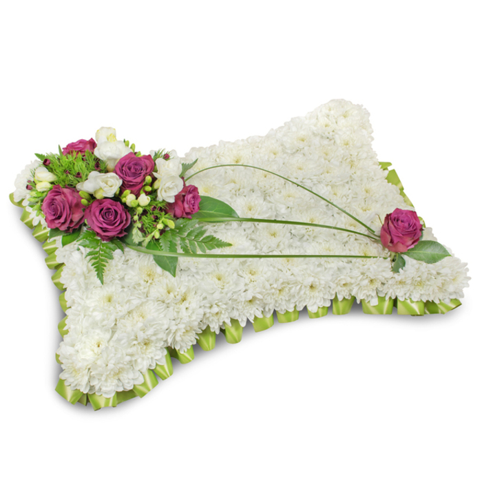 This classic pillow-style funeral arrangement is photographed on a stark white background so the floral forms read with clarity: a plush, rectangular cushion entirely covered in pristine white chrysanthemum florets that create a uniform, serene surface reminiscent of a soft, restful shroud. At the top-left, a composed cluster of deep fuchsia-magentas-velvety roses opened to show layered petals-anchors the piece, punctuated by smaller white freesia or lisianthus that bring delicate contrast and a faint sweet fragrance. Surrounding greenery, including airy fern fronds and fresh green leaves, frames the accent and provides depth against the chrysanthemum field. From the principal cluster, two or three long, dark green bear grass stems sweep diagonally across the pillow, carving an elegant line that culminates in a solitary fuchsia rose placed toward the lower-right, mirroring the main bouquet and completing the visual balance. The edge of the cushion is trimmed with folded light-green satin ribbon, pleated into a scalloped border whose gentle sheen defines the silhouette and lends a refined finish. The composition conveys repose and reverence-soft textures, balanced colour, and thoughtful mechanics that speak to experienced floral construction. I picture this piece being chosen by families in Stanmore and Harrow for a funeral or memorial, its restrained palette and tender accents offering a calm, consoling presence at a service or graveside tribute. For local clients seeking careful handiwork, this pillow embodies quiet dignity and the language of sympathy.