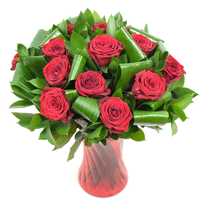 A carefully composed, high-impact floral design centered on a dozen-plus deep crimson roses, each bloom fully open to reveal velvety, tightly spiralled petals that graduate from intense scarlet at the edges to a deeper, wine-coloured centre. The roses are interspersed with plentiful, varied foliage: smaller, dark-green flat leaves create a lush, textured backdrop while larger, glossy leaves - likely aspidistra - are folded into cylindrical and looped forms to produce striking architectural shapes that cradle and frame the rose heads. Those sculpted green elements add volume and structured movement, guiding the eye in soft arcs around the bouquet and offering an appealing contrast in texture and sheen to the soft matte of the petals. The stems are presented in a tall, translucent ruby-red glass vase featuring a subtle vertical rib or twist motif; the vase's narrow waist and gently flared mouth provide balance and a classical silhouette that strengthens the overall composition. Set against a pure white background under soft, diffused studio lighting, the arrangement's colours and textures are rendered with crisp clarity: each petal edge, leaf fold and glass reflection is visible, reinforcing the bouquet's sense of freshness and considered design. This arrangement is ideal for anniversaries, Valentine's Day, or as a luxurious romantic gift - something I might prepare for delivery to a home on Stanmore Broadway or a surprise left at the door near Stanmore Common, where the confident, elegant language of red roses is understood and appreciated.
