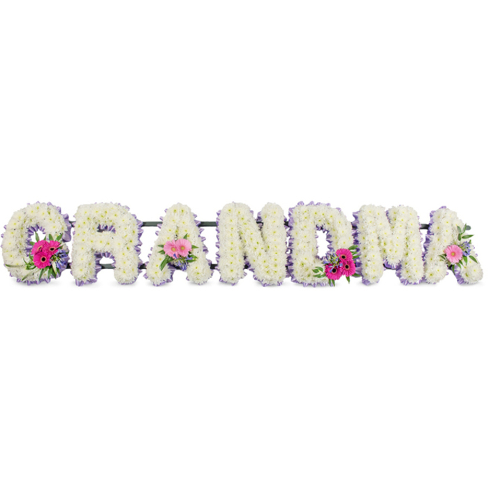In this touching sympathy arrangement, the word GRANDMA is written in flowers, each letter thickly covered with snowy white chrysanthemums that create a cushion-like effect. The blossoms lie close together, their petals layered in gentle waves so the surface appears soft and comforting, almost like a quilt of petals. Running along the outline of every letter is a fine edge of lilac ribbon, the light pastel tone providing a subtle halo around the white flowers and giving the tribute a composed, elegant finish. Positioned thoughtfully along the length of the word are several small, rounded posies: open-faced pink gerberas, tender pink roses just beginning to unfurl, and short stems of lush green foliage. These details introduce warmth and movement, balancing the calm white background with pockets of joyful colour that recall laughter, stories, and treasured afternoons with a grandmother. The lighting is quiet and even, highlighting the texture of the chrysanthemums and the soft sheen of the ribbon without creating harsh contrasts. Imagined in the context of Stanmore, this arrangement might be seen on its way to a quiet service near Stanmore Hill or resting in a chapel close to Canons Park, a floral expression of family love and respect. The tribute feels handcrafted and considered, every bloom placed to ensure clarity of the letters and fullness of the design. Altogether, it offers a gentle, graceful way to say farewell, allowing flowers to convey tenderness, gratitude, and enduring remembrance.