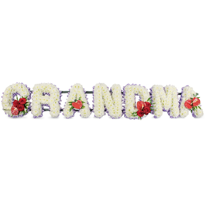 A refined and expressive floral arrangement spells out GRANDMA in bold, three-dimensional block letters, the kind of tribute you might see resting beside a coffin during a quiet service in a Stanmore chapel. Each of the six letters is completely covered with snowy white cushion chrysanthemums, their small, layered petals overlapping to form a dense, cushion-like texture that looks soft yet structured. Just beyond the blooms, a slim outline of pale lilac or soft mauve ribbon can be glimpsed, gently tracing the silhouette of each letter and lending a hint of colour that feels soothing and serene. Throughout the arrangement, rich touches of red inject emotion and depth: on the G, at the far left, sits a cluster of deep red roses accompanied by a striking red-and-white variegated calla lily; on the first A, a single heart-shaped red anthurium is positioned near the base, its glossy, waxy surface contrasted against the matte petals. In the middle, the M is similarly decorated with a group of velvety red roses and another red anthurium, while the final A is adorned with one last anthurium, echoing the earlier details and providing a gentle sense of balance from start to finish. The letters are raised slightly on a dark green metal framework, the sort used by experienced florists like those at Flower Delivery Stanmore when preparing funeral tributes for local churches, crematoria, and cemeteries around Stanmore and Canons Park. Together, the pure white base, the soft lilac outline, and the rich red accents create a tribute that speaks of love, respect, and enduring memory for a cherished grandmother.