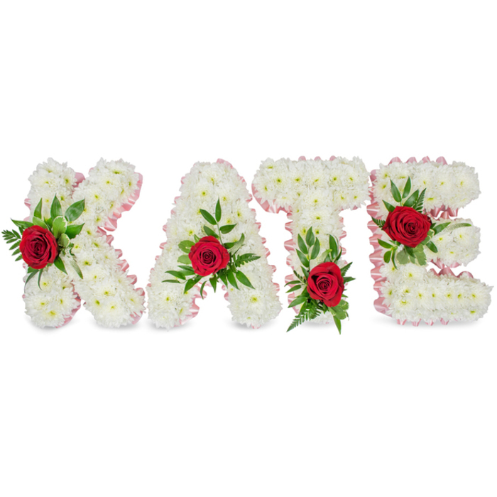 An artisan floral tribute reads plainly and beautifully: KATE formed in large, three-dimensional block letters constructed entirely from dense, pristine white chrysanthemum blooms. The chrysanthemums are arranged so closely that their rounded petals produce a soft, cloud-like surface, conveying a quiet purity and a tactile, almost velvety impression. Along the outline of each letter a fine pale pink satin ribbon is applied with neat pleating, lending a refined border that warms the white and speaks of careful finishing. Each letter features one vivid red rose placed as a deliberate focal point - the roses' deep crimson provides a striking visual counterpoint while their subtle scent would lend a gentle presence in the room. Surrounding each rose is a generous spray of lush green foliage, from broad glossy leaves to feathery ferns, which adds sculptural depth and frames the roses as if cradled. The design is balanced: the K carries its rose and foliage low on the left vertical element, the A is accented at its triangular crown, the T has its red focal bloom near the base of the left half of the crossbar, and the E's highlight sits prominently on the center horizontal bar. Positioned against a clean white background, the arrangement conveys formality and tenderness - the classical palette of white chrysanthemums, pale pink ribbon, red roses and rich greenery is especially fitting for a funeral setting or a memorial display at a local venue such as a Stanmore chapel or community hall. The overall effect is respectful, personalised, and quietly arresting.