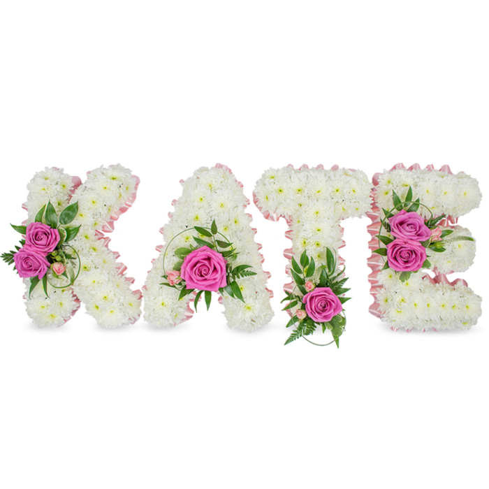 A deeply personal memorial piece, this floral tribute spells KATE in large, sculpted letters made entirely from hundreds of immaculate white chrysanthemums, clustered so closely they form a seamless, cloud-like face to each letter. The inner contours of every character are traced with a pale-pink satin ribbon that imparts a refined, almost handwritten softness to the otherwise bold block forms. Strategic clusters of hot-pink roses provide poignant focal points: two roses adorn the left limb of the K amid glossy green leaves and slender spiral grass; the A holds a single, fully opened rose low in its hollow, framed by delicate pink spray flowers and curling greenery; the T is anchored with a solitary vivid rose at its base, set against ferny fronds; and the E is graced by two roses on its right side with variegated foliage and tiny pink accents woven through. The arrangement is freestanding, photographed against a clean white backdrop with subtle shadows that give it depth and presence, as though ready for placement at a service. The white chrysanthemums offer a soft visual silence while the bright roses bring warmth and personal colour; a faint rose scent mingles with the green freshness of ruscus and eucalyptus beneath. Crafted with the care of a local Stanmore florist, this tribute - prepared by Flower Delivery Stanmore for memorials around Canons Park, Stanmore High Street or Bentley Priory - expresses remembrance, respect and a tender celebration of a life well loved.