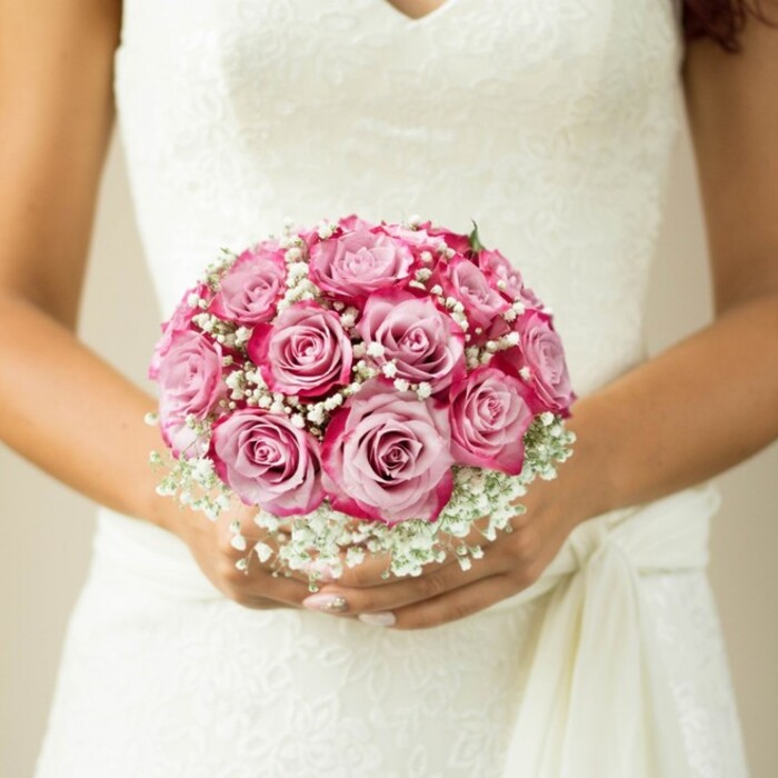 In this evocative wedding image from Stanmore, one can admire the meticulous artistry that goes into the perfect bridal bouquet. Central to the composition, a round arrangement of roses stuns with its striking colour transitions-each bloom glowing with a soft pink core, flaring out to passionate magenta-hued tips. Billowy clusters of baby's breath encircle the roses, imparting classic softness and complementing the bespoke floral lace woven across the bodice of the sleeveless gown. The bride's lightly sun-brushed hands, nails adorned with a soft and tasteful polish, gently support the bouquet above a flowing sash that elegantly knots at the waist. The serene neutral backdrop-a canvas of muted beige-serves only to enhance the intricate floral presentation. Every detail is chosen and arranged with the polished eye of a Stanmore florist, reflecting both local tradition and individual dreams. This bouquet is more than a visual delight; it is a celebration of Stanmore's enduring charm and an invitation to imagine moments of joy along the High Street or beneath the canopy of Stanmore Country Park.