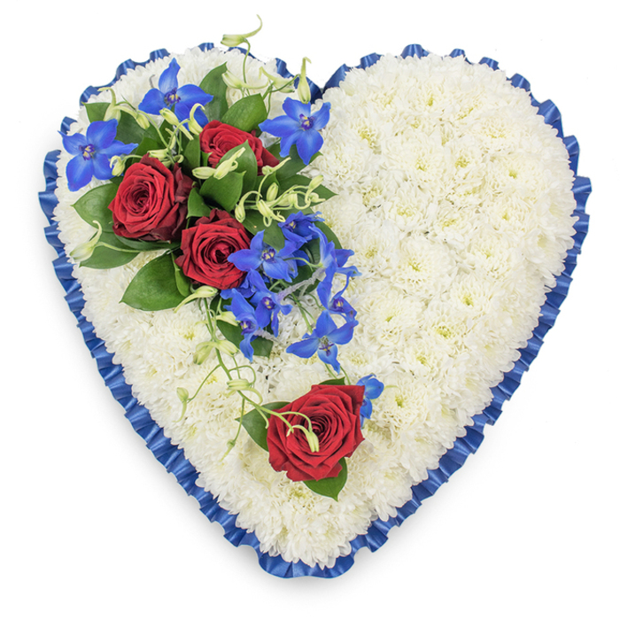 Seen from above, this heart-shaped sympathy arrangement appears like a gentle, floral embrace laid upon a white background, every element thoughtfully placed. The main body of the heart is composed of compact white chrysanthemums, each floret tight and pristine, creating a dense, padded surface that evokes comfort and serenity. Encircling this bed of white is a royal blue satin ribbon, finely pleated and slightly ruffled, which follows the contours of the heart and gives it a defined, ceremonial frame. The satin catches the light in soft highlights and shadows, lending depth and a sense of reverence often associated with memorial tributes in Stanmore's quiet residential streets and green spaces. At the upper left side of the heart, a vivid spray of flowers commands attention. Three fully opened red roses, rich and velvety, rest amidst dark, glossy leaves, their spiralling petals symbolising enduring love and heartfelt goodbye. From this cluster, electric blue dendrobium orchids extend gracefully across the white chrysanthemums, their star-like flowers glowing in intense shades of blue and indigo. Intertwined among the orchids are slender, curling green tendrils, adding softness and a feeling of natural movement, as though the flowers are gently trailing in a breeze. On the lower right curve of the heart, a single red rose has been placed apart, accompanied by a touch of greenery, echoing the passion of the main spray while giving visual balance to the design. Together, the white, red, and blue elements create a powerful yet peaceful tribute, an ideal floral heart for a funeral service or wake in Stanmore, where families gather to honour a loved one and share quiet moments of remembrance.