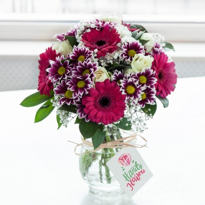 A beautifully composed mixed bouquet sits in a low, clear glass vase, its surface faintly textured so it catches the soft, diffused light like ripples of water. The scene is set against a bright, almost luminous white background that feels like a sunlit window in a Stanmore townhouse, gently blurring away any distractions to keep the attention on the flowers. Dominating the arrangement are several large gerbera daisies in saturated fuchsia and magenta, their wide, daisy-like faces and deep, dark centres creating a bold, confident rhythm throughout the design. Intertwined with them are bicolour chrysanthemums, each bloom a small work of art: outer petals edged in deep purple, fading delicately toward white, and wrapped around golden-yellow centres that echo the warmth of afternoon light over Stanmore Recreation Ground. Tucked between these statement flowers are clusters of creamy white spray roses, some in full bloom with layered, velvety petals, others still in half-open buds that hint at longevity and unfolding emotion. Airy sprays of white baby's breath (gypsophila) weave through the bouquet like mist, softening the strong colours and adding a sense of lightness and volume. Rich green leaves curve and overlap at the base, suggesting freshly cut stems and careful, professional arranging by a local florist who understands how to balance texture and tone. The vase is cinched at the neck with a length of rustic twine tied into a simple, unpretentious bow. From it hangs a crisp white gift tag, decorated with a stylised pink rose motif and flowing coloured script, quietly indicating a handwritten message inside. The overall impression is joyful yet refined, ideal for same-day delivery by Flower Delivery Stanmore to celebrate birthdays, congratulations, or just-because surprises anywhere from Stanmore Broadway to the leafy roads near Belmont Circle.