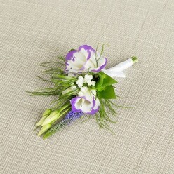 A refined floral boutonniere lies thoughtfully positioned on a pale beige, linen-like fabric, the subtle weave of the cloth creating a calm, neutral setting that allows each flower to be clearly defined. The focal point is a pair of lisianthus blooms, their petals a luminous white with intricate ruffled edges brushed in a saturated purple tone, as though hand-painted with a fine artist's brush. Nestled in around these main flowers are tiny white fillers reminiscent of baby's breath, forming delicate, cloud-soft clusters that lend volume while still keeping the design airy and light. Rising through the centre, a slender stem of muscari, or grape hyacinth, displays its miniature bell-shaped blossoms in a rich, blue-violet hue, adding both height and a gentle woodland charm. Wisps of fine, feathery greenery curve naturally around the flowers, adding texture and the sense of something freshly cut from a local Stanmore garden after a passing shower. Two small, pale green buds, still tightly closed, sit quietly within the composition, symbolising moments yet to unfold on a significant day. The stems are gathered together with care and wrapped in a smooth white satin ribbon, finished with a simple bow that looks ready to be secured to a suit jacket. The overall impression is one of understated sophistication, ideal for a groom, best man, or prom guest arriving at a Stanmore venue near the Broadway or along Marsh Lane. It conveys that blend of personal attention and timeless style that local florists bring to intimate celebrations and formal events alike.