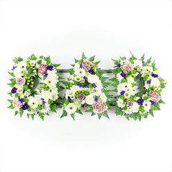 This image shows a touching floral tribute spelling DAD in three solid, capital letters, laid out horizontally against a spotless white background that draws attention to every floral detail. The letters are formed from a densely packed bed of white chrysanthemums and crisp white gerbera-like blooms, creating a plush, textured surface that suggests softness and quiet strength. Small white buds and dainty filler flowers fill any gaps, giving the impression of a continuous blanket of white, symbolising purity and peace. The shapes of the letters are outlined and softened with lush green foliage, especially fern fronds and tidy greenery that echo the natural calm of Stanmore's leafy streets and green spaces. Bright green button chrysanthemums or small clustered blooms are tucked among the leaves, adding a lively, fresh contrast that brings the tribute gently to life. Across this white and green base, the florist has placed clusters of deep purple and rich violet flowers - similar to pansies or spray chrysanthemums - that lend depth, dignity, and a hint of formality, befitting a funeral or graveside service. These stronger tones are balanced by soft lavender and pale mauve roses and frilled carnations, their petals opening in layers that feel tender and affectionate. The lighting is bright yet gentle, with no distractions behind the arrangement, as though the tribute has been carefully positioned on a studio table before being delivered to a local chapel or home in Stanmore. The overall effect is of a lovingly crafted sympathy design, a traditional yet bespoke DAD arrangement that speaks clearly of love, loss, and cherished memories.