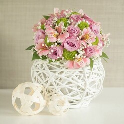 Drawing on Stanmore's peaceful charm, this floral centerpiece invites both touch and admiration-a showcase of meticulous craftsmanship and garden-fresh beauty. The arrangement rises from a distinctive white vase, its bulbous form and elaborate, open lattice reminiscent of bespoke pottery found in local Stanmore studios. Within, a lush and rounded bouquet bursts with full-bloom pastel pink and lavender roses, their velvety petals soft to the gaze. Pink alstroemeria provide elegant drama with their signature striping and subtle throats of yellow, while zesty pops of lime-green button chrysanthemums add playful contrast and modern verve. Wisps of white baby's breath drift between blooms, infusing the display with an ethereal lightness akin to dawn mist in Canons Park. Interwoven dark green leaves lend depth and a grounding earthiness. To one side, a pair of decorative spheres crafted from pale rattan or bamboo strips-one substantial, the other smaller-mirror the creative texture of the vase, evoking the tactile spirit celebrated throughout Stanmore's home-grown craft fairs. Bathed in gentle light, the entire tableau rests upon a spotless white surface, set before a softly textured beige wall that accentuates every pastel hue. The effect is both welcoming and sophisticated, perfect for marking birthdays, anniversaries, or wedding table arrangements across Stanmore, and capturing the area's quiet joy and creative heart.