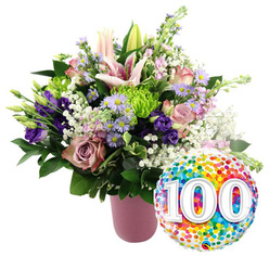 This image shows a joyful 100th celebration arrangement set against a clear white background, the kind of display you might send to a much-loved grandparent in Stanmore. The focal point is a lush bouquet arranged in a stylish, ribbed vase finished in a soft, matte pink, its gentle tone echoing the romance of the blooms. Emerging from the vase is a full, rounded composition of flowers in shades of blush, mauve, purple, lime green, and white. Light pink Oriental lilies rise elegantly from the centre, some blossoms fully open to reveal their star-shaped petals and long stamens, others held in elongated buds that add vitality and movement. Nestled among them are velvety roses in soft pink and deeper dusty rose hues, their layered petals unfolding like a well-told story. Deep purple lisianthus provide a dramatic contrast, their ruffled forms adding depth. Scattered throughout are delicate lavender asters with sunny yellow centres, evoking wildflower borders you might find near Stanmore Country Park. Lime green button chrysanthemums offer cheerful pops of colour and tactile, cushion-like textures. Wisps of white baby's breath drift through the bouquet, softening the edges and lending an airy, cloud-like effect. Glossy dark green foliage encircles the arrangement, grounding the pastel shades and creating a sense of fullness. On the lower right side, a shiny round foil balloon completes the composition. Its metallic surface is covered in colourful confetti-style dots-bright reds, oranges, yellows, greens, blues, and purples-whirling around large, bold white numbers "100" with a slight shadow that gives them a raised appearance. The entire scene feels festive yet elegant, perfectly suited to celebrate a 100th birthday, a centenary wedding anniversary, or a hundred years of memories for families in and around Stanmore.