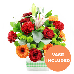Captured in bright, even light against a white background, this arrangement presents a joyful mix of colours and textures in a low rectangular glass vase encircled with a soft green and white gingham ribbon, a detail that brings to mind the gentle, suburban charm of Stanmore's tree-lined lanes. The bouquet feels plentiful and uplifting, its flowers gathered closely together in a painterly composition. Deep red roses, with densely layered, velvety petals, offer a classic romantic note, while nearby red and red-orange carnations bring softness and volume with their delicately frilled edges. Interspersed throughout, bold orange gerbera daisies with dark central disks stand like small, sunlit faces, their clean, symmetrical petals radiating vibrancy reminiscent of a sunny afternoon in Stanmore Country Park. Counterbalancing these warm tones, clusters of lime-green button chrysanthemums appear as small, round cushions of colour, adding a crisp, modern twist and a lively sense of rhythm to the design. Rising from within the bouquet, white Oriental lilies add height and elegance; several blooms are open wide, revealing subtle pink stripes and golden stamens, while tall buds wait to open over the coming days, extending the life and changing character of the display once delivered. Around the blooms, a generous collar of glossy dark green foliage and fine, silver-green eucalyptus leaves gives structure, depth, and a natural, garden-inspired feel, as if the flowers were freshly cut from a well-tended Stanmore garden. Through the clear glass, the submerged stems create a clean, linear counterpoint to the softness above. In the bottom right corner of the image, a bright orange circular label reads "VASE INCLUDED" in bold white capitals, indicating that this vibrant bouquet is supplied complete and ready to place straight onto a table or reception desk.