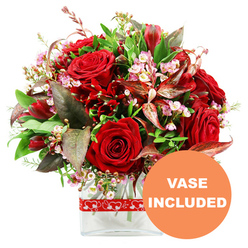 An elegant, passion-filled floral arrangement rests in a clear glass vase, set against a crisp white background that highlights every detail, much like a bouquet ready to go out from a Stanmore florist's workbench. The focal point is a cluster of velvety red roses, each bloom at a different stage, from tightly furled buds that feel full of anticipation to more open flowers revealing their layered hearts. Weaved amongst them are smaller, equally rich deep red flowers, reminiscent of alstroemeria or similar multi-petalled stems, building texture and giving the eye plenty to explore. Threaded delicately through the red tones are sprays of tiny, soft pink and creamy white waxflowers or limonium, their petite blossoms forming clouds of airy lightness, softening the intensity of the colour palette. A lush frame of greenery surrounds the blooms: wide, glossy green leaves, finer stems of lighter foliage, and distinctive burgundy and reddish-brown leaves with subtle veining that echo the warmth of the roses and add an extra layer of sophistication. The bouquet is shaped into a generous, rounded dome, suggesting a hand-tied style crafted with care and precision. It is anchored in a simple, modern rectangular glass vase, the stems arranged cleanly in the clear water, giving the design a contemporary, uncluttered feel. A vivid red ribbon, decorated with a repeating motif of white hearts, encircles the vase, reinforcing the bouquet's romantic intention-perfect for an anniversary dinner near Stanmore Hill or a surprise delivery to a home close to Stanmore Country Park. In the lower right corner of the image, a strong orange circular overlay bears the text "VASE INCLUDED" in neat white uppercase letters, signalling that the arrangement arrives complete and ready to display the moment it is received.