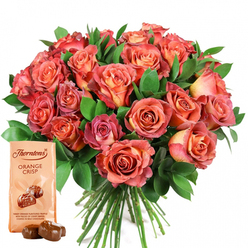From the experienced hands of a Stanmore florist comes this evocative bouquet-roses in harmonious shades ranging from fiery coral through soft peach to rich salmon pink, their petals opening in gentle cascades to form a lush, almost glowing mound. Each rose seems to capture a different shade of a sunset watched from the rolling hills near Stanmore Country Park, with their creamy-yellow centers gradually fading into deep, velvety rims. Framing this brilliant display, broad leafy greenery-satin-surfaced and deep green-offers fresh contrast and amplifies the bouquet's warmth. The bouquet is accentuated by a thoughtfully placed pouch of Thorntons Orange Crisp chocolates: its light beige wrapping stands out with sophisticated branding and a depiction of glossy chocolate truffles, tempting both the eyes and the palate. Before the packet, two milk chocolate truffles rest, their sheen inviting curiosity about the crispy wafer and zesty orange within. Every detail-from the interplay of colour and texture to the soft glow of petals in natural light-embodies a sense of celebration, tenderness, and indulgence. Against the clean white background, the full bouquet and artisan confectionery form a harmonious tableau, ideal for elevating birthdays, anniversaries, or simply bringing heartfelt cheer to loved ones in and around Stanmore, especially along Marsh Lane and the serene surrounds of Bentley Priory.