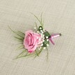 A single pink rose boutonniere is displayed from a gently elevated viewpoint, resting on a softly textured, light beige linen-style fabric that feels very much like the relaxed, natural textiles you might see in a Stanmore townhouse near Canons Park. The rose itself is a fresh, semi-open bloom in a blush-to-rose-pink gradient, its velvety petals curving outward in layered spirals, with the palest pastel pink at the heart slowly deepening to a richer, more romantic tone at the edges. Nestled closely around the base of the rose are petite clusters of white baby's breath, each tiny, cloud-like flower forming a delicate halo of softness that contrasts with the more substantial rose petals. Fine, wispy strands of bright green, grass-like foliage fan gently from behind the bloom, giving the piece a light, airy movement, while a broader, deeper green leaf is neatly folded to cradle the stems, adding structure and a touch of tailored polish. The stems are bound with a lustrous satin ribbon in tones of lavender-pink and deeper magenta, wrapped smoothly and tied in a compact knot that feels refined rather than fussy. Soft, natural light falls across the boutonniere, picking up the texture of the linen background and the subtle sheen of the ribbon, creating a calm, romantic mood that would suit a wedding at a local Stanmore venue or a prom near Stanmore station. The overall effect is one of timeless elegance: a small, handcrafted floral accent designed to sit perfectly on a lapel, radiating gentle colour, freshness, and quiet celebration throughout the day.