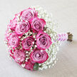 A hand-tied bouquet of roses rests on a pale, off-white fabric that appears softly textured, like fine table linen or the hem of a bridal veil spread across a dressing table in Stanmore. The arrangement lies on a gentle diagonal, with the flower heads fanning out to the upper left and the bound stems pointing to the lower right. Around a dozen roses form the heart of the bouquet, their petals unfurling in layered gradients of pink. At the centre of each bloom, the colour is a subtle, dusty mauve and lavender-pink, gradually intensifying into brighter fuchsia and deep magenta towards the outermost petals. Many of the roses are semi-open, revealing tight swirls of overlapping petals that create a sense of richness and texture, as if each flower contains its own secret. Cloaking the roses is a generous collar of white baby's breath, or gypsophila, its tiny blossoms scattered so abundantly that they resemble a gentle mist or a ring of lace encircling the stronger pink tones. Occasional glimpses of deep green leaves peek from beneath, anchoring the design and hinting at its garden origins. The stems are trimmed and brought together at the lower right, bound with a smooth, lilac satin ribbon that glows faintly in the soft light. A slender strand of small white pearls winds around this ribbon, adding a sense of bridal refinement and ceremony. The atmosphere is calm, intimate, and full of promise, perfectly suited to a wedding day in Stanmore, a renewal of vows at a local synagogue or church, or an anniversary celebration at home, with the bouquet reflecting the kind of thoughtful craftsmanship available for local flower delivery across Stanmore and nearby neighbourhoods.