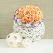 Beneath diffuse daylight this arrangement reads like a small sculpture: roughly two to three dozen apricot and soft peach roses arranged in a compact dome, their petals layered and tactile, each blossom giving off a gentle, honeyed fragrance. White Lisianthus ring the composition, their creamy faces delicately edged in deep violet - an unexpected, dramatic flourish that frames the roses and draws the eye around the bouquet. Interspersed throughout, tiny clusters of white Gypsophila (baby's breath) introduce a whisper-light texture that keeps the mass feeling buoyant rather than heavy. The entire bouquet is set inside a contemporary rounded white woven vase, an open lattice of reed or rattan painted bright white, whose artisanal pattern contrasts beautifully with the roses' softness. On the tabletop to the left, two woven wooden spheres - one larger, one smaller - repeat the basket's geometry and add an organic, decorative rhythm. A soft, blurred linen-toned wall behind the display keeps the focus on colour and form. This arrangement carries a refined yet approachable mood: romantic enough for a wedding centrepiece, warm enough as an anniversary gift, and modern enough to lift a Stanmore sitting room near Stanmore Broadway or the Jubilee Line station. For locals seeking same-day delivery within Stanmore and nearby Harrow, its balanced textures, scent, and palette make it an immediately comforting, polished choice for celebrating new beginnings or quiet milestones.