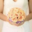 This arrangement carries a soft, poetic hush suited to a bridal moment, where each apricot rose unfolds like a small, warm sun. The roses are gathered in a symmetrical cluster, their outer petals curling with creamy texture and apricot glow, while a neat halo of purple-tipped lisianthus circles the core, adding a cool, twilight accent to the composition. A delicate lattice of white baby's breath threads through the flowers, lending an airy, weightless quality as though the bouquet might lift on a summer breeze. The lighting is gentle and even, brushing the petals to reveal faint veins and a subtle sheen that suggests freshness and careful handling. Against the bride's gown-be it antique lace or modern satin-this bouquet reads as both timeless and current, the kind of piece that photographs softly and feels intimate in the hand. The designers who work in Stanmore often place such a bouquet in ceremonies that begin near Stanmore Station or unfurl across the greens of Stanmore Common; here the floral language is restrained but lush, written in peach and mauve rather than loud colour. Available for local delivery through Flower Delivery Stanmore, this bouquet is suited to couples seeking understatement with emotional depth: a compact, refined assembly that supports the day rather than dominating it, and that lingers in memory as a quiet centrepiece of joy and promise.