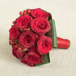 A lush dome of red roses rests on a pale beige, softly textured cloth, the simplicity of the background allowing the bouquet's intensity to shine. The roses are arranged in a neat, rounded cluster, forming a compact yet abundant shape that feels both classic and contemporary. Each bloom is open and velvety, its petals layered tightly into intricate spirals. The colours drift from bright scarlet at the outer edges to deeper, more wine-red tones within, giving the impression of depth and emotion. A few petals gently loosen and curl, suggesting that the bouquet has been freshly handled, perhaps just carried through the streets of Stanmore from a ceremony near Belmont Circle to a reception by Canons Drive. Nestled between the blooms are polished clusters of red hypericum berries, their glossy, spherical forms sitting slightly above the petals and catching light in tiny reflections, adding a crisp, tactile contrast to the softness of the roses. Around the base, broad dark green leaves are folded with precision, overlapping to create a smooth collar that frames and supports the design. The stems disappear beneath a band of vivid red satin ribbon that wraps the handle visible to the right, its sheen creating a subtle highlight that mirrors the luxurious feel of the flowers. Gentle, diffuse lighting enhances the velvety texture of the petals, the sleek shine of the berries, and the faint weave in the background fabric. The arrangement feels undeniably romantic, perfect for a Stanmore bride, a special anniversary dinner near Stanmore Hill, or a heartfelt gesture delivered to a family home off The Broadway.