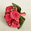 A compact, luxurious bouquet of deep red roses and berries rests on a soft, light beige fabric, the gentle texture of the background setting off every detail of the flowers. Around five or six velvety roses, each in full bloom, form the vibrant heart of the arrangement, their petals layered in rich shades of crimson and scarlet that suggest warmth, romance, and timeless elegance. Threaded between the roses, small clusters of rounded Hypericum berries in reddish-pink and red-orange tones add playful contrast, their smooth surfaces catching the light and bringing a lively sparkle to the design. Broad, dark green tropical leaves are folded and curled with a florist's precision, cupping the flowers in a lush frame that feels both sculptural and natural. A neatly tied red satin ribbon secures the stems at one side, its sheen echoing the depth of the petals and hinting at careful, hand-tied craftsmanship. The lighting is soft and even, like a calm afternoon in a Stanmore townhouse, illuminating each texture without harsh shadows. The bouquet could be imagined as a boutonnière-style piece for a modern groom, an intimate bridal accessory, or a special gift for a romantic evening at home after a walk through Stanmore Country Park. Every element speaks of considered design and fresh, carefully selected stems, the kind of piece you might have delivered locally for a small but meaningful celebration in HA7.