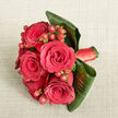 This image shows a sophisticated, compact bouquet resting on a light beige, linen-like fabric, the soft texture of the background allowing every floral detail to stand out. Viewed from above, the focal point is a tight cluster of five to six deep red roses, each bloom fully open and lush. Their petals are thick and velvety, arranged in graceful spirals that naturally draw the eye towards their dark, inviting centres, evoking the feeling of an intimate celebration in Stanmore's quiet residential streets. Slipped between the roses are polished clusters of small hypericum berries, round and bright red with a faint orange undertone. Their glossy skins catch the light, offering a lively counterpoint to the matte softness of the petals and suggesting cheerful festivity, as if made for an anniversary toast or engagement dinner near Canons Drive. Surrounding the flowers is a collar of broad, dark green tropical leaves-likely cordyline or ti-curved and wrapped expertly around the base to frame the bouquet. These leaves provide structure and a contemporary silhouette, their subtle sheen contrasting beautifully with both the berries and the roses. Around the stems, a coordinated deep red satin ribbon is wrapped neatly, its smooth, reflective finish showing just beneath the leaves and completing the design with a sense of tailored elegance. The lighting is gentle and evenly spread, bringing out the richness of the reds and the depth of the greens while keeping the overall mood calm and romantic. This bouquet feels ideal for a Valentine's Day surprise delivered across Stanmore, a proposal dinner near Bentley Priory, or as a chic wedding-party bouquet carried down the aisle in one of the area's local venues.