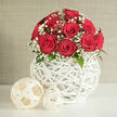 A compact, domed arrangement of vivid red roses sits at the centre of a serene scene, every bloom carefully angled so that the velvety spirals of petals are clearly visible. The roses are rich and saturated in colour, forming a rounded canopy that looks soft yet structured, as if crafted specifically for a romantic occasion. Between them, threads of white baby's breath emerge, the tiny round blossoms creating a gossamer veil that lightens the intensity of the crimson. Just beneath this halo of blooms, glimpses of dark green leaves peek out, polished and tidy, providing a subtle transition into the vessel below. The bouquet is set within a sculptural, spherical vase made of slender white strips woven into an airy lattice, reminiscent of contemporary wicker art. Through its open design, the neutral background slips in, while soft daylight filters through, casting faint, intersecting shadows on the pristine white tabletop. Beside the vase, two woven spheres in a warm cream hue lie side by side, one larger and one smaller, mirroring the texture and craftsmanship of the vase while gently softening the composition. Behind everything, a muted beige-grey wall creates a calm, modern backdrop similar to stylish interiors found around Stanmore, perhaps near Stanmore Country Park or the quiet roads leading towards Canons Park. The lighting is smooth and even, without harsh contrasts, bringing out every texture-from the plush rose petals to the fine strands of the woven ornaments. The overall impression is one of understated elegance and heartfelt sentiment, ideal for a wedding display in Stanmore, an anniversary gift, or a refined statement piece for a special dinner at home.