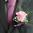 This image shows a delicate pink rose boutonniere resting against a charcoal-grey suit jacket, the kind of classic finishing touch often chosen for weddings in Stanmore and the surrounding leafy streets. The rose is a soft, romantic pink, its petals gently unfurling in elegant spirals, each edge smooth and velvety, as if carefully checked and selected by an experienced florist. Tiny clusters of white gypsophila-baby's breath-nestle around the bloom, creating a light, cloud-like halo that enhances the feeling of tenderness and new beginnings. Rising behind the central flower, fine, feathery greenery adds height and a natural, slightly wild texture, contrasting beautifully with the crisp, structured fabric of the lapel. At the base of the arrangement, a lilac satin ribbon wraps tightly around the stems, tied neatly so that the ribbon's gentle sheen catches the light, mirroring the sheen of a special-occasion tie. The overall colour palette-blush pink, snowy white, cool green, and lilac-feels softly celebratory, perfectly suited to a ceremony in Stanmore's churches, synagogues, or nearby country venues. The ambient light is calm and flattering, allowing every detail to be seen: the curve of each petal, the tiny star shapes in the gypsophila, the fine grain of the suit cloth. It suggests a moment just before the event begins, perhaps a final adjustment in the mirror before stepping out to greet guests. There is a quiet intimacy in this small floral accent, as if it embodies gratitude, love, and the gentle pride of taking part in a milestone day that will be remembered long after the rose has passed its prime.