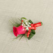 Crafted with the eye of a seasoned florist, this boutonniere centres on a fierce magenta rose whose tightly cupped petals promise bloom and warmth. Surrounding the bloom, clouds of delicate white baby's breath soften the composition and lend a vintage whisper, while a trio of shiny red hypericum berries nestle close, offering punctuation and a festive glint. Fine, feathery green grasses sweep outward like a breeze through late-summer hedgerows, counterbalanced by wider, sculpted leaves folded to create a tailored frame around the focal flower. The stems are wrapped in a confident band of crimson satin ribbon, wound neatly and finished with a small bow that reads as both practical and decorative. The arrangement rests diagonally on a light beige, subtly woven fabric surface, the neutral tone and gentle texture acting as a quiet stage for the vivid colours and tactile contrasts. You can almost feel the velvety petal edges and the smooth hardness of the berries, and the scent is a soft murmur of rose tempered by fresh greenery. This boutonniere suits joyous milestones-weddings at nearby Canons Park halls, prom nights leaving from Stanmore station, or anniversary dinners in the village. As a Stanmore-based florist familiar with local weddings and fêtes, I value the piece's blend of spirited colour and considered restraint; it delivers presence without overpowering, and it pictures well against both a morning suit and a summer blazer.