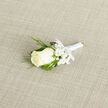 Set on a subtly textured light beige weave, this boutonniere features a single, immaculate creamy white rosebud that commands the eye with its velvety, tightly layered petals. Encircling the bud are tiny clusters of pure white gypsophila-baby's breath that reads like a soft, cloudlike halo-while narrow, feathery sprigs of dark green foliage, suggestive of cedar or evergreen, lend a slightly woodsy depth. Broader, deep green leaves provide a sculpted backdrop and ensure the rose remains the focal point. The stems are carefully gathered and wrapped in a smooth white satin ribbon, tied in a small, deliberate bow that reflects the same quiet care found in every detail of the piece. The boutonniere is placed on a gentle diagonal, catching the light in a way that emphasizes contrasts between the creamy bloom, the crisp white accents, and the rich greens. There is a restrained, ceremonial fragrance implied-the faint, fresh scent of rose layered over cool green notes-evoking a calm morning before vows. Crafted with the knowledge of a long-practised hand, this accessory is intended to be pinned close to the lapel as a symbol of purity and new beginnings. Flower Delivery Stanmore often prepares pieces like this for couples marrying near Bentley Priory or exchanging vows on Stanmore High Street, and the neutral background here highlights the fine textures and makes the boutonniere a quietly elegant choice for formal events across the local area.