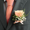 There is a practiced restraint in this boutonniere's design-a single flawless peach rose set against a charcoal lapel, its blush tones melting into coral-pink highlights that echo a carefully chosen silk tie. The rose's petals are velvety to the eye and imply a soft, sweet scent; surrounding it, sprays of tiny white baby's breath form a delicate, frothy cloud, lending a light, whimsical texture as if conjured from early-morning mist over Stanmore Common. Slender green foliage weaves through the composition, giving subtle support and a fresh, dewy note reminiscent of local cuttings gathered near Cannons Park. At the base, a neat satin ribbon in matching coral ties everything together in a small, tidy bow whose sheen catches the light like a promise. The charcoal suit serves as a refined canvas, deepening the floral tones so the boutonniere reads clearly in photographs taken along Stanmore High Street outside the chapel or on the steps of a family venue. Prepared with the care of an experienced local florist, this boutonniere from Flower Delivery Stanmore is designed to feel both personal and ceremonial-ideal for a groom, best man, or honoured guest seeking an elegant, understated floral accent for a Stanmore wedding or celebratory gathering.