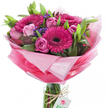A joyful, hand-tied bouquet captured against a brilliant white backdrop, the composition centring on several large gerbera daisies in a vivid fuchsia pink, each bloom marked by a deep, almost black centre that makes the petals glow. Interwoven among the gerberas are multiple delicate pink roses in varying stages of bloom - some open with soft, layered petals, others held as taut, promising buds - their softer pastel tones balancing the gerberas' intensity. Small clusters of deep purple-blue filler flowers, likely statice, thread through the arrangement, lending fine texture and a subtle cool contrast to the warm pinks. Lush green foliage, from broad glossy leaves to slender stems, shapes a verdant backdrop that gives the bouquet volume and a sense of natural depth. The flowers are gathered and wrapped in a crinkled, sheer pink cellophane that graduates from an almost translucent pastel halo at the top to a richer pink toward the base, the wrap catching light and adding a celebratory sheen. The bundled stems are visible through a clear cylindrical glass vase that holds the design upright; a simple length of rustic twine is tied around the vase neck and suspends a small white rectangular tag bearing a stylised pink-and-green rose logo, a quiet sign of careful floristry. The scent is gently sweet and fresh, like a morning market stall on Stanmore Broadway, and the overall mood reads as bright, affectionate, and perfect for birthdays, Mother's Day, or to brighten a room near Canons Park or Stanmore Common. An arrangement like this feels personal and hand-crafted, made to convey warmth and well-wishing to someone close.