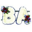 A quietly powerful memorial arrangement forms the capital letters BA, each letter built from hundreds of small, pristine white chrysanthemums packed close to create a pillowy, cloud-like surface. The letters' contours are crisply defined with a ruffled royal blue satin ribbon that frames the white blooms and gives each character a dignified, sculpted edge. At the upper left curve of the B sits a rich cluster of deep red roses - some tightly furled, others softening into open petals - paired with spikes of intense blue delphinium-like flowers and glossy dark green leaves; a mirrored cluster of the same red roses and electric blue florets nestles into the lower right of the A, with pale green trailing tendrils that soften the lines. The whole pair rests side-by-side on a low, dark green rectangular base that stabilises the tribute and adds a subtle grounding tone against the clean white backdrop. Under soft studio light the chrysanthemums read as pure and peaceful, while the red and blue accents punctuate the composition with notes of love, courage, and solemnity. As a florist who works in Stanmore, I picture this tribute placed at a service near Stanmore High Street or Bentley Priory, the colour palette echoing quiet dignity in a chapel or community hall. Texturally the piece combines the dense, matte softness of the chrysanthemum heads with the velvety roses and ribbon sheen, and there is a faint rose scent suggested by the red blooms. This bespoke letter tribute is a considered, personal way to honour a life - a visual message of remembrance, respect and gentle farewell tailored for local memorials in the Stanmore area.