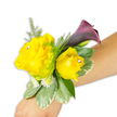 A vibrant wrist corsage is photographed diagonally across a light-to-medium forearm against a crisp white background, the composition designed to show how the piece is worn. The focal pair of buttery yellow roses-one larger and fully open on the left, the other a slightly smaller, more cupped bloom to the right-carry tiny clear rhinestones nestled at their centers that catch the light like miniature stars. Behind and slightly above the roses a single, smooth calla lily unfurls in a dark plum to nearly black-cherry hue, its trumpet form providing a sleek, dramatic counterpoint to the soft rose petals. Around these main blooms a florist's hand is evident in the textural layering: feathery asparagus-fern filigree lifts and lightens the silhouette, while clusters of tiny white filler blooms-resembling spray chrysanthemums or delicate baby's breath-add fullness and a whisper of sweetness. Broad, variegated leaves with creamy edges (pittosporum-like foliage) form a stable base, their waxy surfaces grounding the arrangement and echoing the polished feel of the rhinestones. The corsage is secured by a narrow band that is mostly concealed beneath the flowers so the wrist appears naturally adorned. You can almost sense the soft rose scent and the cool, satiny feel of the calla's petal against skin. Perfect for prom, a bridesmaid or a guest of honour, this joyful yellow-and-plum palette suits celebrations across Stanmore-from a photoshoot on Stanmore High Street to a reception near Canons Park-where local florists often craft similar wearable pieces for special moments.